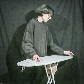 Noura stands behind an ironing board, in one hand is a red stethoscope handle on the ironing board, in the other hand is a transducer on the ironing board.